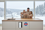 Lade das Bild in den Galerie-Viewer, Familie mit Kinder sitzt glücklich in der Küche & trinkt Wasser mit Blick aus dem Fenster auf Winterlandschaft in der Küche steht ein Wasserfilter 12 cm schmal weiß , liegt unter der Küche versteckt liegt auf dem Boden klein & kompakt eingebaut und heißt Smart Quelle 12 centimeter hoch - mit Aufschrift groß Produkt des Monats mit 55% Rabatt