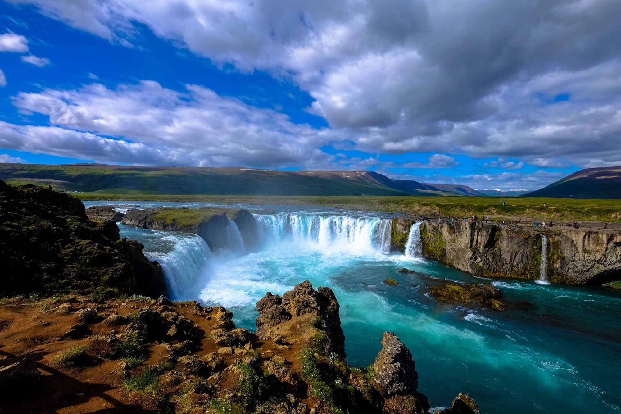 Breiter Wasserfall in offener Landschaft, darunter ein türkisfarbener Fluss